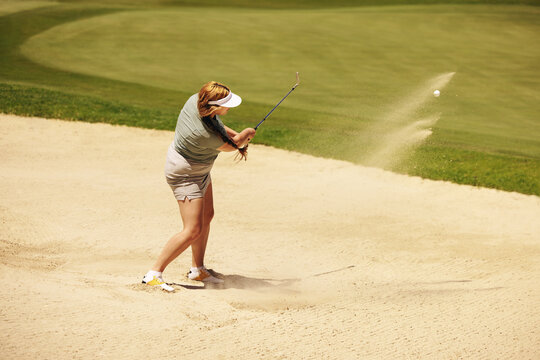 Female amateur golfer skillfully hits golf ball from bunker during a sunny day on the course, demonstrating precision and focus in her game