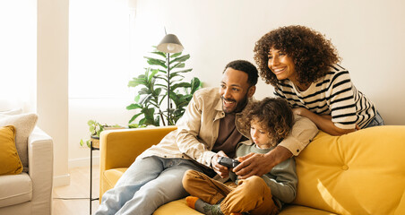 Smiling parents and their young child enjoying a bonding moment on a bright sofa, playing video games together in a warm and cheerful living room at home.