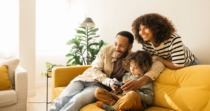 Smiling parents and their young child enjoying a bonding moment on a bright sofa, playing video games together in a warm and cheerful living room at home.