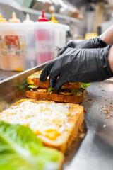 A close-up of hands wearing black gloves assembling a sandwich in a kitchen. Fresh ingredients include lettuce, pickles, and cheese on toasted bread, showcasing a delicious meal preparation.