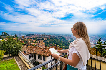 City break in Italy. Beautiful mature woman tourist standing and holding city map at viewpoint on autumn day. Panorama of city Bergamo. Sightseeing. Side view