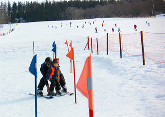 Caucasion father teaching his 4-year old boy how to ski in the baby ski park. Little skier with a knitted hat and a warning vest going downhill slalom with his father in the ski school.