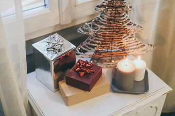 Gift boxes near lighted candles and Christmas tree inside home. Box with holiday ribbon with bows...