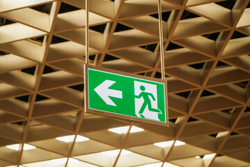 A green emergency exit sign hangs from the ceiling of a building. The sign displays a left arrow and a running figure, indicating the direction of an exit route.