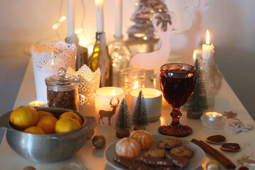 Various Christmas decorations, cookies, chocolate, nuts and tangerines, wine glasses and lit candles on the table. Holiday hygge at home. Selective focus.
