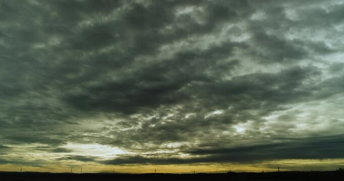 Gray Cloud Deck Racing Toward the Camera Over a Dark Flat Horizon Lined with Power Pole Silhouettes While a Narrow Band of Yellow Glow Sits Just Above the Landscape in a Moody 4K Time Lapse