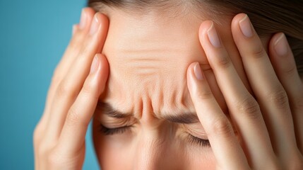 Fototapeta premium Woman experiences headache while holding her head with both hands against a blue background in bright light.
