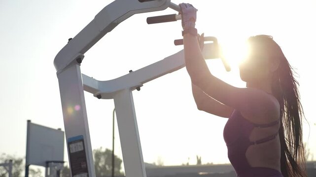 The silhouette of a slender woman with long dark hair on a pull-up simulator on an outdoor sports field. A woman trains outside.