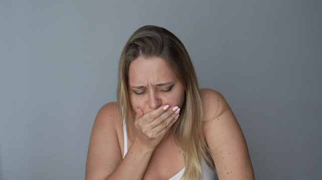 Young blonde woman covering her mouth and holding her chest with hand because of the nausea on a dark grey background. Morning sickness, toxicosis, heartburn, poisoning, eating disorder
