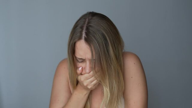 Young caucasian blonde woman covering her mouth and holding her chest with hand because of the nausea on a dark grey background. Morning sickness, toxicosis, heartburn, poisoning, eating disorder