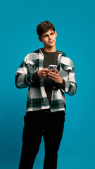 Portrait of young handsome man in casual clothes holding smartphone and texting message with contemplation against blue studio background. Concept of youth, emotions, lifestyle, communication