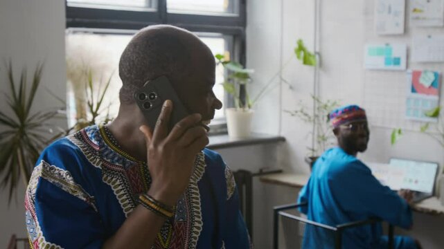 Bald Black man in patterned blue African shirt talking to colleague and smiling, while taking phone call and working at computer in bright open space office with plants and wall charts