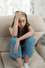Young woman holding her head, frustrated, sitting on sofa in modern living room during daytime