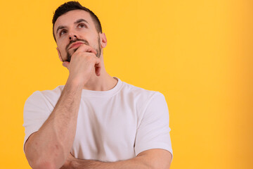 A young man is standing against a bright yellow background, looking up thoughtfully with a hand on...