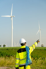 Vertical image of back of wind turbine or windmill engineer worker point to his right side with row of wind turbine in the background.