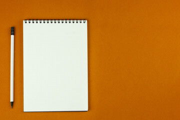 A notebook with an even spring at the top with white pages and a white pencil with a gray lead and a black eraser lie on an orange background on the left with space for text. High quality photo