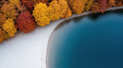 A stunning aerial shot of a lake fringed by trees in vibrant autumn hues, forming a natural border between the tranquil water and rocky shore.