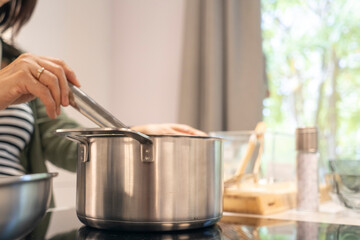 Young woman is cooking in kitchen, holding utensil and preparing food with pot