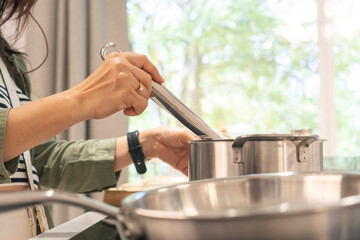Young woman is cooking in kitchen, holding utensil and preparing food with pot