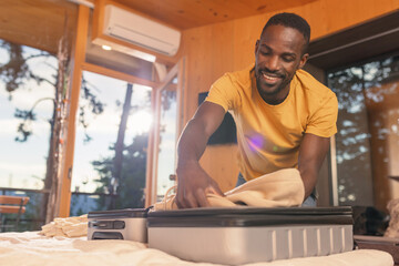Smiling African American man packing his suitcase in a bedroom, preparing for travel, a...