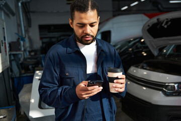 A handsome young mechanic reviews his phone while savoring a warm drink in the garage.