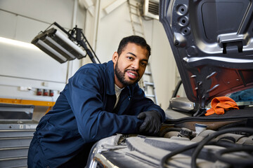 A skilled young mechanic smiles while inspecting an engine under a bright garage light