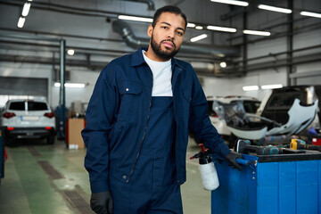 Focused mechanic in blue work attire posing confidently in a busy auto repair shop