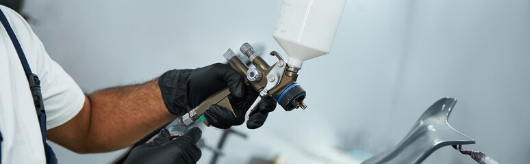 Handsome mechanic skillfully uses spray gun to paint a vehicle in a bright garage space.