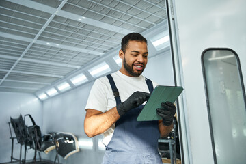 Handsome young mechanic reviews job details on a tablet in the busy workshop during daylight.
