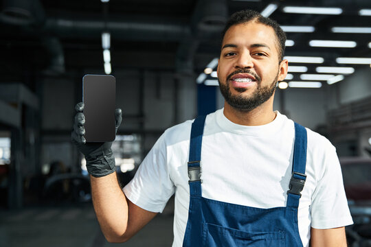Handsome young mechanic smiles while showcasing a smartphone in a bright workshop setting. - Powered by Adobe