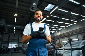 Young mechanic in blue overalls uses smartphone while surrounded by vehicles in workshop.