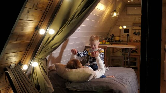 Mother and son have fun lying on the bed in a small wooden triangular house, they play and cuddle. Vacation in a country house in autumn. View through the window with flashlights and a thick curtain.