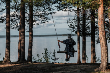 A woman is swinging on a tree swing in a forest near a lake. The scene is peaceful and serene, with the sound of the water and the rustling of the trees in the background