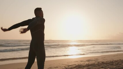 A surfer in a wetsuit stretches on the beach at sunrise, preparing for a surf session. He embraces the serene atmosphere and the beauty of nature.