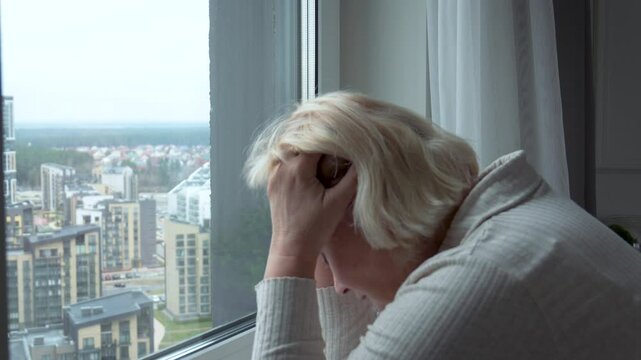 Retired woman feeling anxious while looking through window at city below. Old female worried about future and state of world. Lonely sick elderly person experiencing grief or headache with migraines