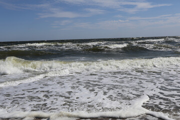 Rolling Waves on a Windy Day: A dynamic view of ocean waves crashing onto the shore under a partially cloudy blue sky, showcasing the power and beauty of nature's energy.
