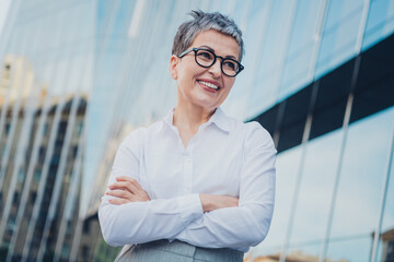 Confident middle-aged businesswoman with short grey hair and glasses outdoors in urban cityscape, wearing elegant white shirt.