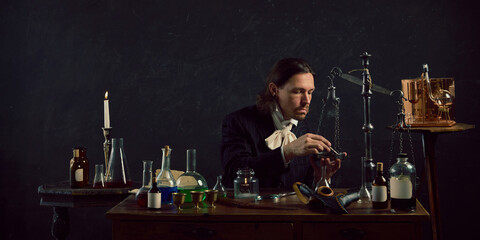 Young man, dressed vintage costume sitting in vintage laboratory and study of ancient remedies against dark mystery background. Concept of comparisons of eras, medicine, science, pharmaceutical. Ad