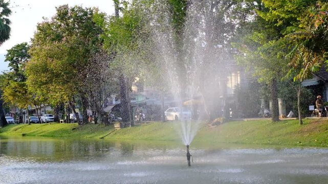 A small fountain is spraying water into a pond. The water is spraying out of a nozzle and is spraying into the air. The pond is surrounded by trees and grass