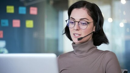 Smiling female customer service representative wearing headset, working on laptop, engaging with clients. Businesswoman conducting virtual meeting, providing support and assistance in office. Close up