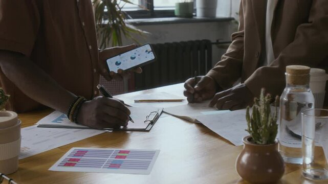 Two unrecognizable African American men working with graphs and documents, analyzing statistics at table in modern eco-friendly office workspace
