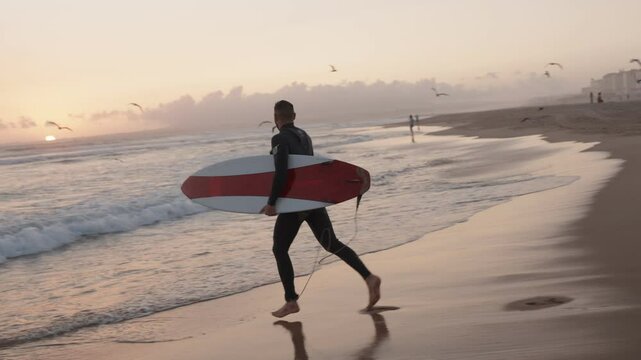 A surfer in a wetsuit runs along the beach at sunset, carrying his surfboard, ready to catch some waves.