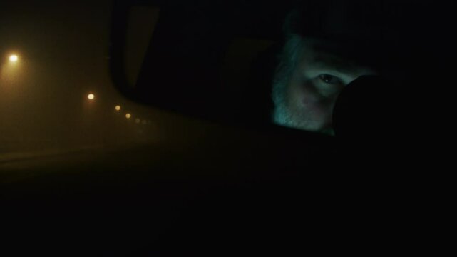 Night shot. Suspicious man, gumshoe, looking at the rear view mirror with a grim expression, inside darkened parked car. portrait, reflection with greenish light on face. High quality 4k footage