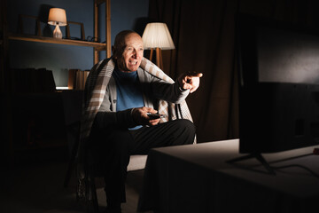 Pensioner with remote control in hand watching comedy or humor channel laughing and pointing finger at screen at night in living room of house