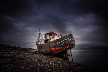 Old shipwreck rests on the shore under a cloudy sky at dusk near a tranquil body of water