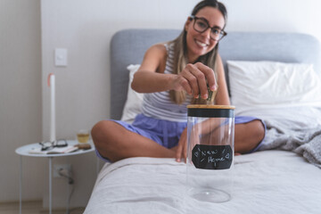 A woman is sitting on a bed and putting coins into a jar. New home concept.