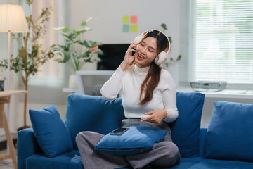 Happy businesswoman enjoying music using wireless headphones and talking on the phone while sitting on comfortable blue sofa in modern living room at home