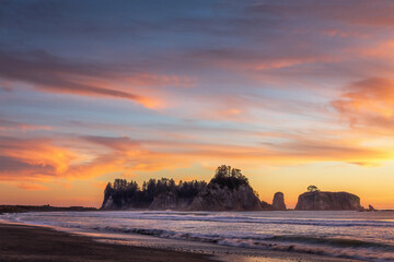 Fototapeta premium La Push beach at Olympic National Park, Washington, during sunset. Waves roll gently onto the shore, surrounded by sea stacks, driftwood, and pebbles, with a warm orange sky creating a peaceful view.