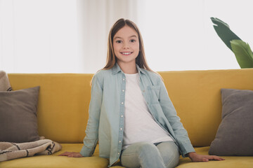 Obraz premium A cheerful young girl with long hair and denim shirt smiling while sitting comfortably on a yellow sofa in a bright living room