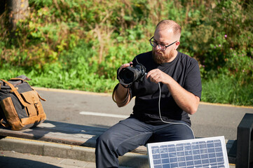 Bearded male photographer sits on bench, connecting camera to solar panel for charging. Integration...